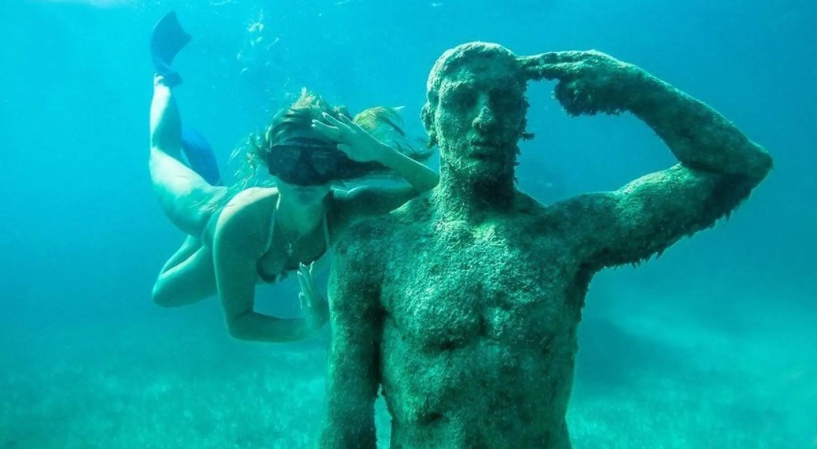 Young snorkeling woman imitating the salute gesture of an underwater man sculpture at Punta Nizuc reef